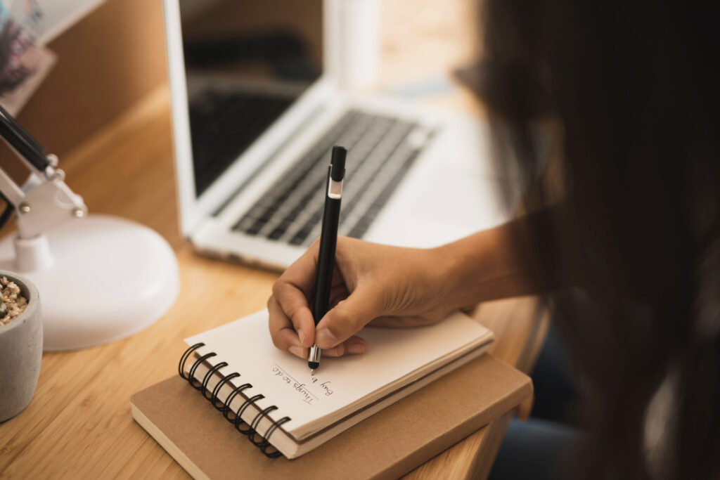 Quais são as etapas e o cronograma do edital para novos polos de pós-graduação Imagem ilustrativa de pessoa anotando em um caderno pautado sobre a mesa, ao lado de notebook aberto e luminária, em referência às etapas previstas no edital.