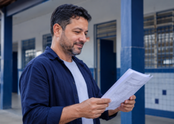 Professor da rede pública analisa documentos em corredor de escola, representando a Rede de Interlocutores do Enem 2026.