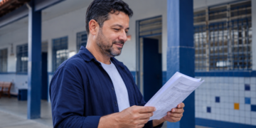 Professor da rede pública analisa documentos em corredor de escola, representando a Rede de Interlocutores do Enem 2026.