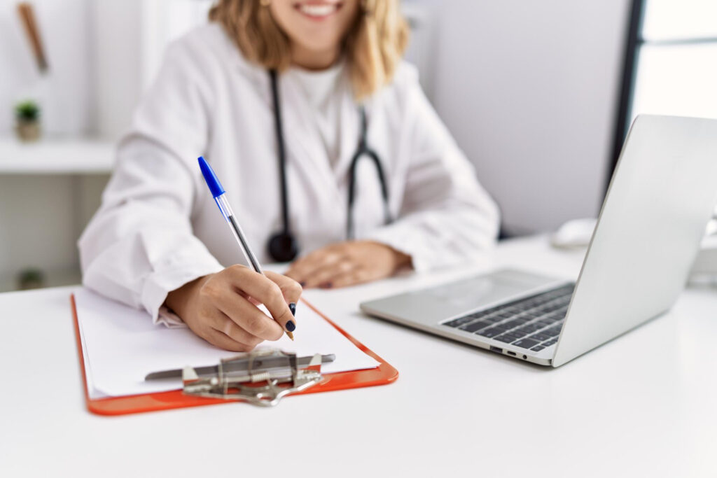Data do resultado definitivo do Enamed e do Revalida 2026/2 Imagem ilustrativa de médica de jaleco branco preenchendo um formulário em prancheta, com notebook aberto sobre a mesa do consultório.