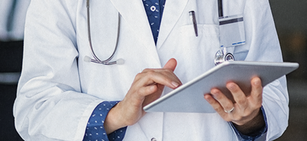 Qual a importância do Enamed Imagem ilustrativa: médico de jaleco branco e estetoscópio no pescoço toca a tela de um tablet, cena que remete à importância do Enamed