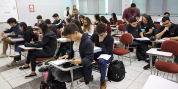 Estudantes sentados em carteiras de uma sala de aula cheia, com cadernos abertos e mochilas no chão, durante atividade de preparação para as provas do Enem 2026.