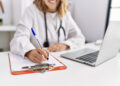 Imagem ilustrativa de médica de jaleco branco sentada à mesa preenchendo um formulário em prancheta, com notebook aberto ao lado.