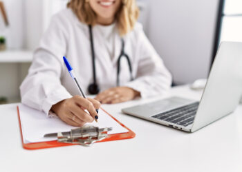 Imagem ilustrativa de médica de jaleco branco sentada à mesa preenchendo um formulário em prancheta, com notebook aberto ao lado.