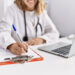 Imagem ilustrativa de médica de jaleco branco sentada à mesa preenchendo um formulário em prancheta, com notebook aberto ao lado.