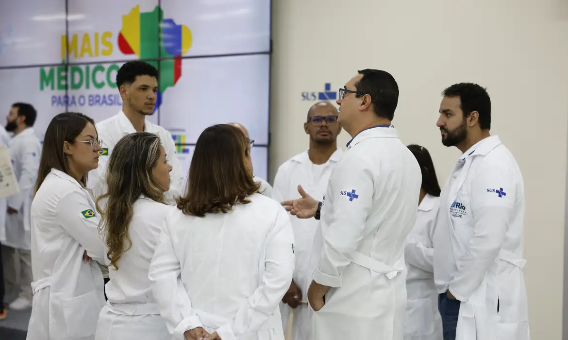 Etapas da avaliação do Revalida para médicos formados no exterior Grupo de médicos de jaleco branco reunido em conversa durante evento institucional do Ministério da Saúde, com painel do programa ao fundo.