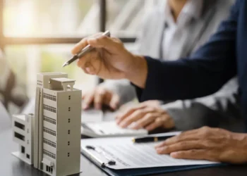 Curso de Administração para Condomínios reúne 80 horas de conteúdo sobre gestão e legislação 5 Profissionais analisam documentos de administração condominial ao lado de maquete de prédio residencial.