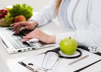 Curso de Nutrição Clínica aborda alimentação, saúde e cuidados nutricionais para diferentes pacientes; saiba como se inscrever 5 Profissional de nutrição utilizando notebook em mesa com frutas, estetoscópio e materiais de atendimento, representando o estudo da Nutrição Clínica.