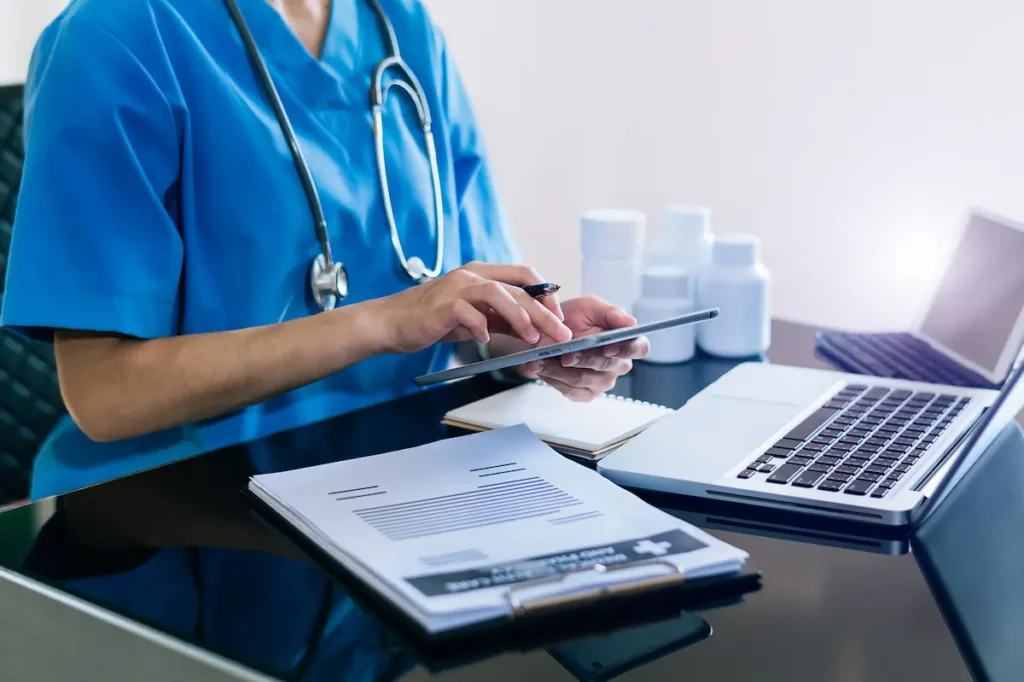 Profissional de saúde utiliza tablet e notebook em atividade ligada à administração hospitalar.