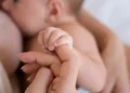 Bebê durante a amamentação segura a mão da mãe em imagem sobre curso de aleitamento materno.
