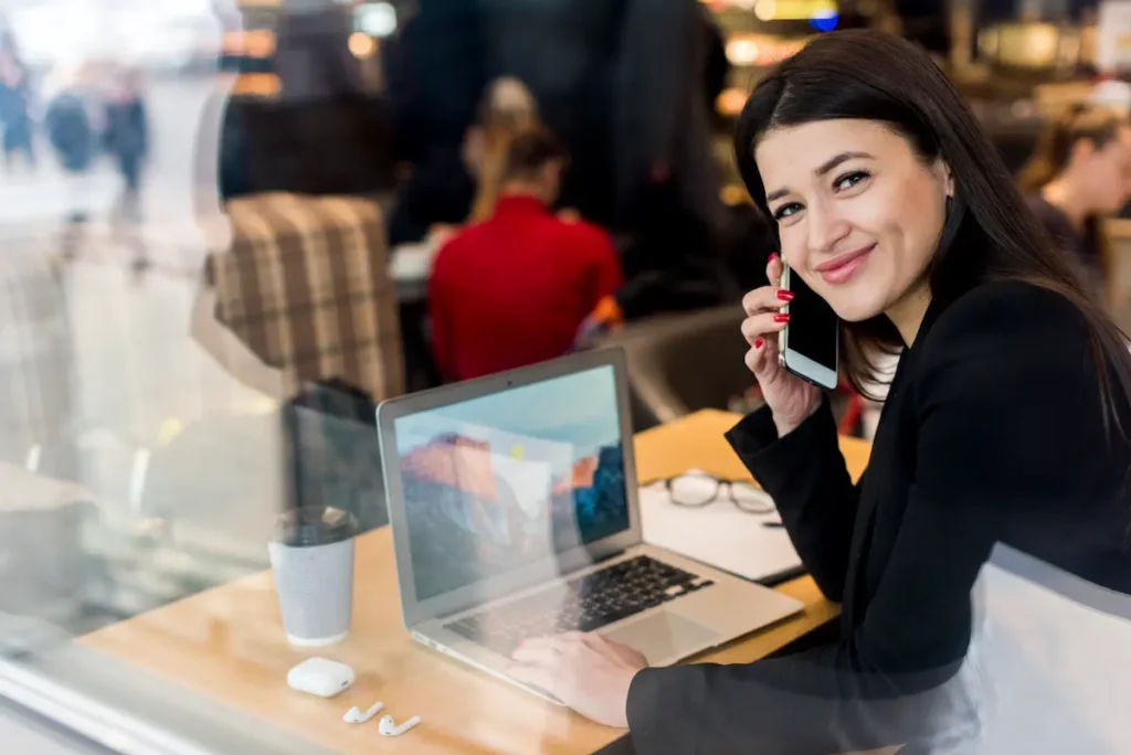 Mulher com celular junto ao ouvido e notebook aberto sobre a mesa em uma cafeteria.