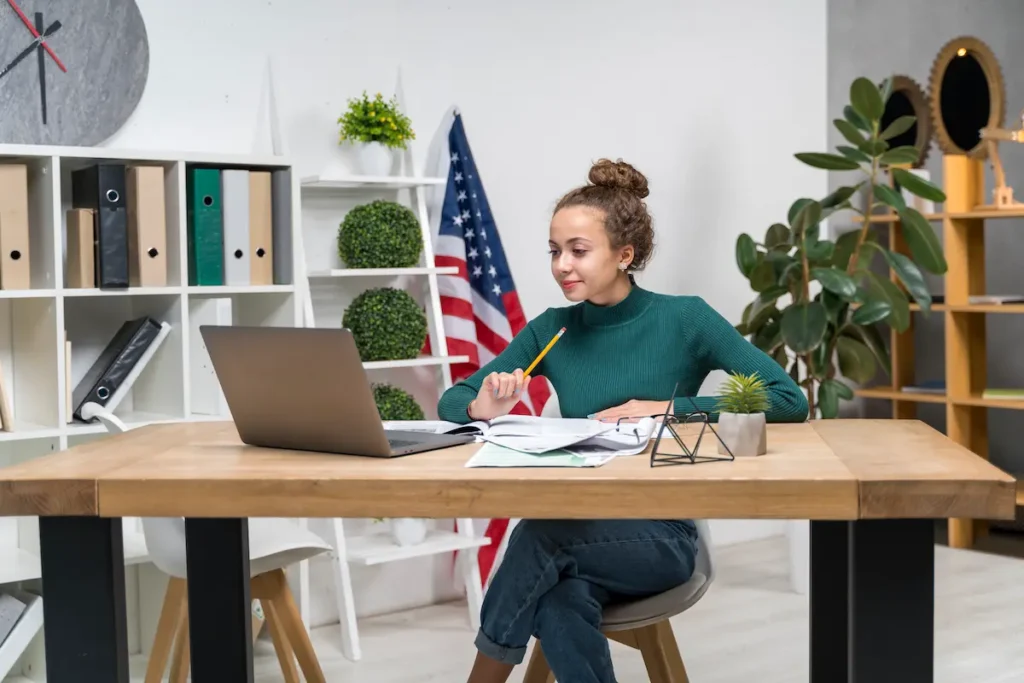 Mulher estudando com notebook e caderno sobre a mesa, com bandeira dos Estados Unidos ao fundo.