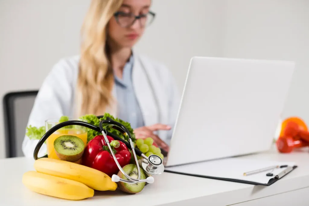 Profissional de nutrição utilizando notebook em consultório, com frutas, vegetais e estetoscópio sobre a mesa, ilustrando o Curso de Nutrição Clínica da Bizu.