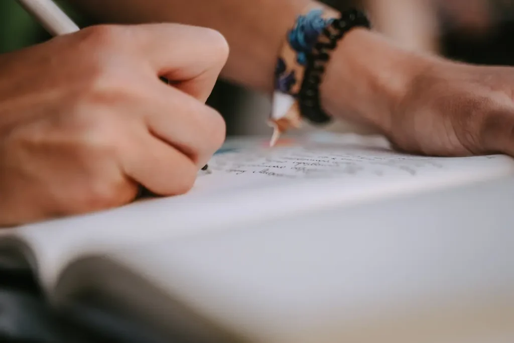 Pessoa escrevendo em um caderno com uma caneta, representando a prática de redação e o desenvolvimento da escrita para vestibulares e concursos públicos.