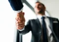 Curso de Técnicas de Vendas online reúne 39 módulos sobre clientes, negociação e estratégias; veja como se inscrever 2 Duas pessoas com roupas sociais apertam as mãos, com foco no cumprimento e um homem desfocado ao fundo.