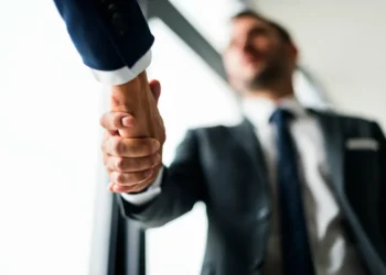 Curso de Técnicas de Vendas online reúne 39 módulos sobre clientes, negociação e estratégias; veja como se inscrever 5 Duas pessoas com roupas sociais apertam as mãos, com foco no cumprimento e um homem desfocado ao fundo.