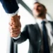 Curso de Técnicas de Vendas online reúne 39 módulos sobre clientes, negociação e estratégias; veja como se inscrever 11 Duas pessoas com roupas sociais apertam as mãos, com foco no cumprimento e um homem desfocado ao fundo.
