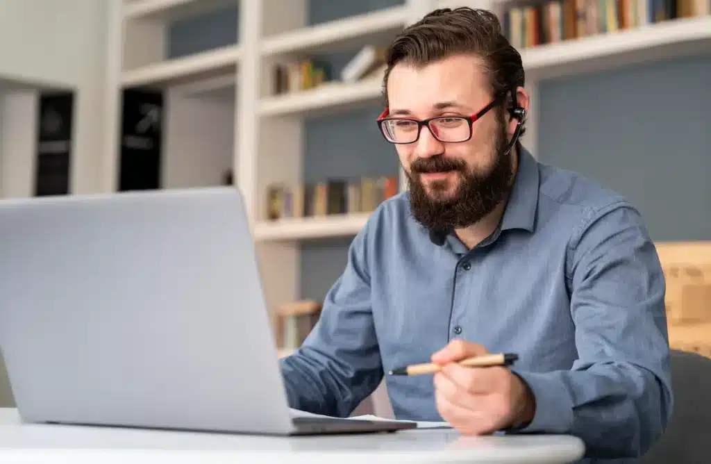 Curso online de Vendas B2B ensina estratégias para conquistar clientes e melhorar negociações 1 Homem com óculos e fone de ouvido utiliza notebook e segura uma caneta diante de uma estante de livros.