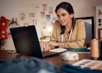 Curso online de Vendas B2B ensina estratégias para conquistar clientes e melhorar negociações 5 Mulher utiliza notebook em um ateliê, com tecidos e materiais de costura sobre a mesa.