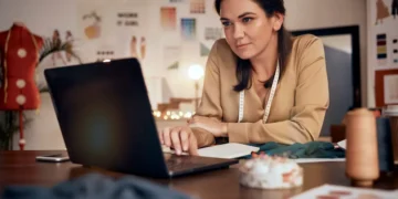 Mulher utiliza notebook em um ateliê, com tecidos e materiais de costura sobre a mesa.