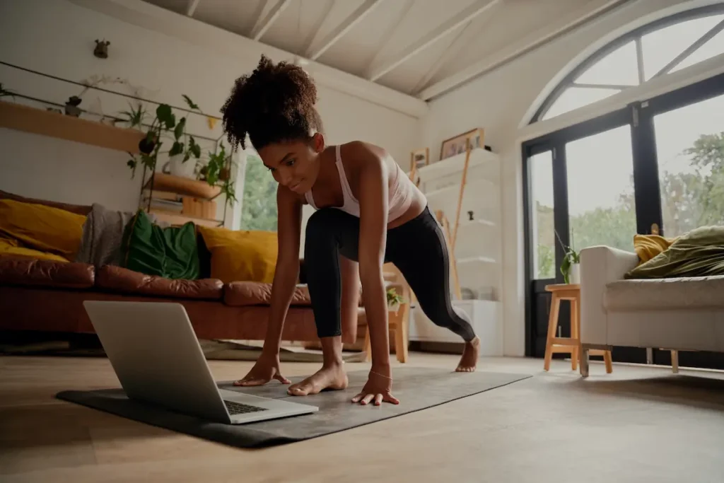 Mulher se exercitando sobre um colchonete na sala de casa, com um notebook à frente e grandes janelas ao fundo.