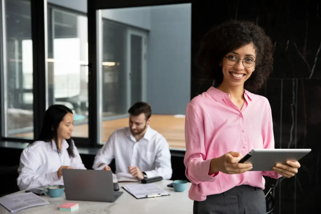 Profissional de Recursos Humanos utilizando um tablet em ambiente corporativo, enquanto colaboradores trabalham em equipe ao fundo.