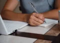 Dificuldade para escrever uma redação? Curso de 80 horas ensina técnicas para melhorar a escrita e desenvolver textos com mais confiança 11 Pessoa escrevendo em um caderno ao lado de um notebook, representando o curso online de Técnicas de Redação da plataforma Bizu.