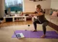 Treinamento funcional em casa: curso online ensina exercícios sem precisar de academia 11 Mulher fazendo agachamento com halteres sobre um tapete na sala de casa, com um notebook à frente.