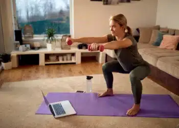 Treinamento funcional em casa: curso online ensina exercícios sem precisar de academia 4 Mulher fazendo agachamento com halteres sobre um tapete na sala de casa, com um notebook à frente.