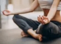Yoga para iniciantes: curso online ensina técnicas de respiração, meditação e posturas básicas 11 Mulher sentada de pernas cruzadas pratica yoga e meditação, com as mãos em posição de mudra, exercitando a concentração e o relaxamento.